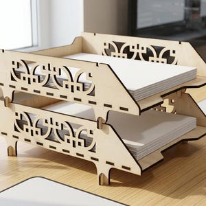 Könnte beinhalten: Eine Dokumentenablage mit zwei Ebenen aus hellem Holz mit lasergeschnittenen geometrischen Mustern an den Seitenteilen. Der stapelbare Organizer hält auf jeder Ebene Stapel von weißem Papier und verfügt über Steckverbindungen.