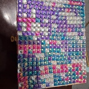 May include: A decorative box covered in small, square, iridescent gems. The gems are arranged in a mosaic pattern, with colors including purple, pink, blue, and white. The box has a gold-colored clasp on the side.