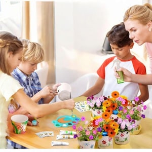 May include: A group of people are gathered around a table, engaged in a painting activity. The table holds paint supplies, including paint tubes, a palette, and paintbrushes. Several flower pots with colorful flowers are also present, suggesting a gardening or craft theme.