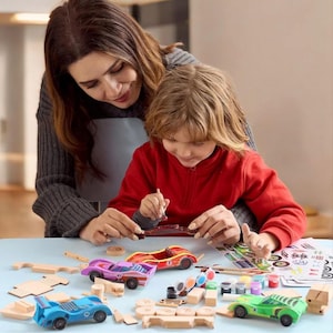 May include: A woman and child are painting wooden toy cars. The cars are in various colors, including blue, purple, red, and green. Paint pots, brushes, and car parts are on a light blue surface, suggesting a craft activity.