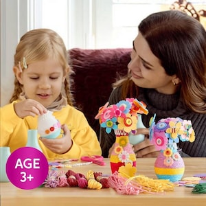 May include: A craft activity set with a young child and an adult. The set includes colorful vases decorated with yarn and felt flowers, along with craft supplies. The image includes the text "AGE 3+".