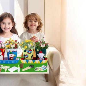 May include: Three colorful flower pots with painted designs of a snowman, a monkey in a Santa hat, and other festive characters. Each pot holds vibrant flowers. The pots sit on a tray with craft supplies, suggesting a DIY project.