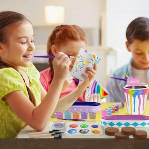 May include: Children painting ceramic pots with colorful paints. The pots are decorated with various designs, including fireworks and dripping paint effects. Paintbrushes, paint trays, and a pot with a "Cosmos" sign are also visible, suggesting a craft activity.