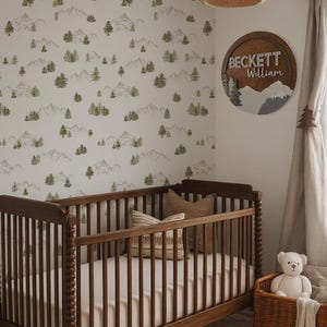 May include: A nursery scene featuring a wooden cot, a wicker basket with a teddy bear, and a rug with a geometric pattern. The walls are decorated with wallpaper depicting mountains and trees. A wooden sign with the name "Beckett William" is displayed.