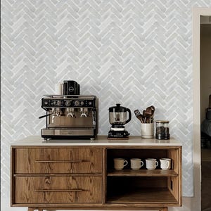 May include: A kitchen scene featuring a wooden cabinet with a white countertop. It holds a professional espresso machine, a coffee maker, and mugs. The background is a light gray herringbone pattern.