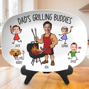 May include: White oval platter with the text "Dad's Grilling Buddies" and cartoon illustrations of a man grilling, children, and a dog. The man is wearing a red apron and holding tongs. The platter is on a black stand.