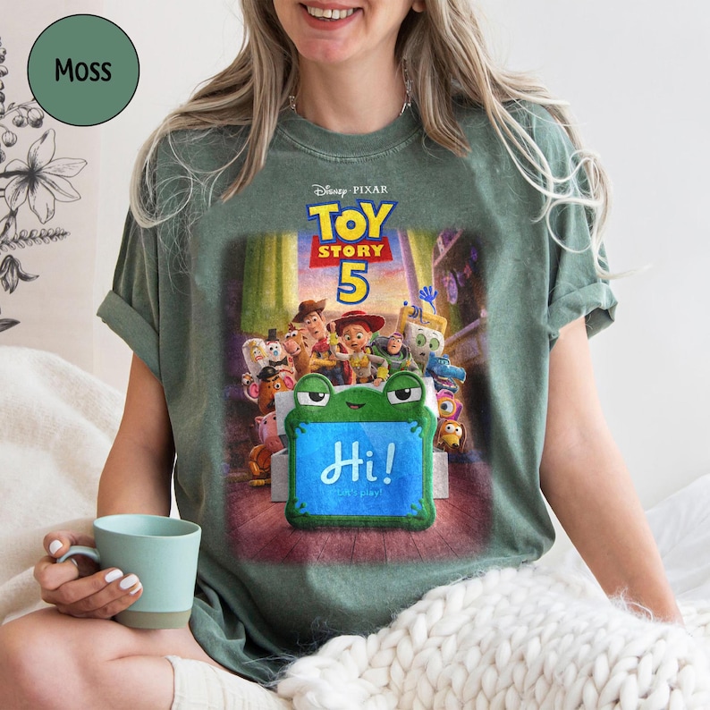 Puede incluir: Camiseta verde musgo con el cartel de la pel&iacute;cula Disney Pixar Toy Story 5. La camiseta tiene un gr&aacute;fico de los personajes y el texto "Hi! Let's play!" en un letrero con forma de rana. La camiseta es de cuello redondo y manga corta.