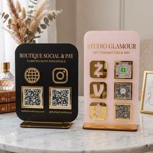Peut inclure: Deux panneaux rectangulaires en acrylique sur supports dorés. Le panneau noir porte les inscriptions BOUTIQUE SOCIAL & PAY, CURATED SHOP EXPERIENCE, thefashiontheory.com et @thefashiontheory. Le panneau rose indique STUDIO GLAMOUR, GET CONNECTED & PAY.