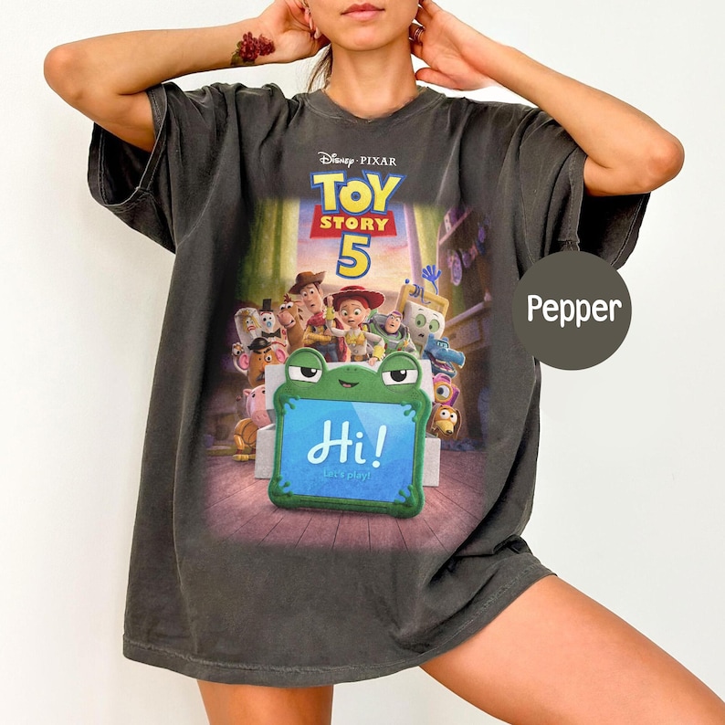 Puede incluir: Camiseta gris oscuro con el p&oacute;ster de la pel&iacute;cula Disney Pixar Toy Story 5. El dise&ntilde;o muestra personajes y un letrero con forma de rana con "Hi!" en azul. La camiseta es de corte holgado.