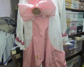 May include: A pink and white dress with a large pink bow and a gold emblem. The dress has a white collar and long sleeves.
