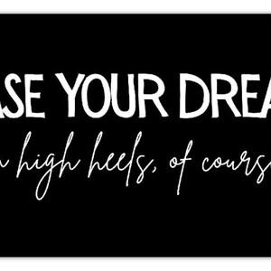 May include: Black rectangular sign with rounded corners. The white text reads "CHASE YOUR DREAMS" above "in high heels, of course." in a cursive font. The sign is a motivational piece.