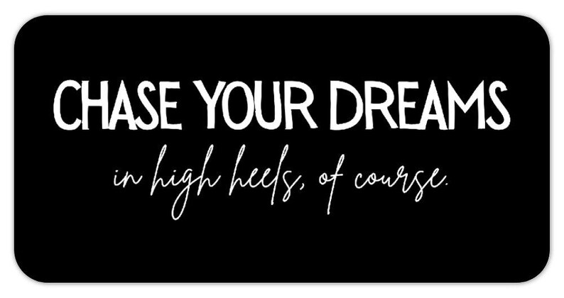 May include: Black rectangular sign with rounded corners. The white text reads "CHASE YOUR DREAMS" above "in high heels, of course." in a cursive font. The sign is a motivational piece.