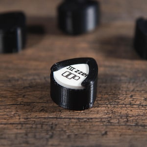 May include: A black 3D-printed guitar pick holder with a white pick inside, featuring the text "Jazz III". Several other black holders are blurred in the background, all on a wooden surface.