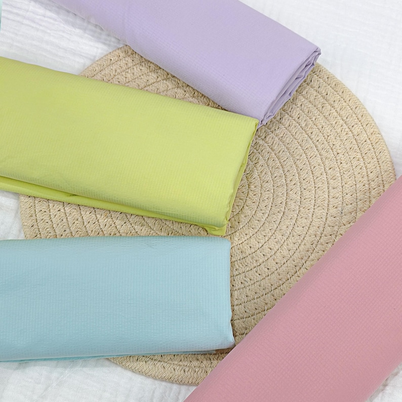 May include: An overhead shot displays folded fabric in pastel colors: light green, light blue, lavender, and pink. The fabric is arranged on a round, woven placemat. The background is a white, textured surface.