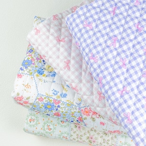 May include: A stack of quilted blankets in various patterns and colours. The top blanket is a light purple gingham with pink bows. Below it is a pink and white gingham blanket, and a floral patchwork blanket. The bottom blanket has a light green floral pattern.