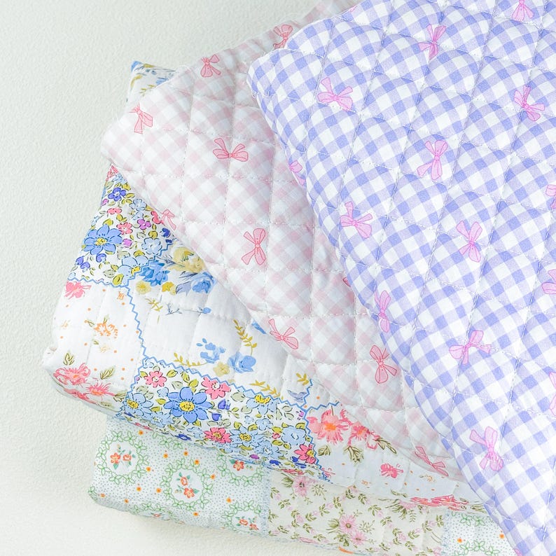 May include: A stack of quilted blankets in various patterns and colors. The top blanket is a light purple gingham with pink bows. Below it is a pink and white gingham blanket, and a floral patchwork blanket. The bottom blanket has a light green floral pattern.