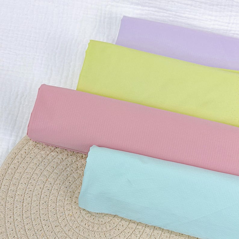 May include: An overhead shot of pastel-colored fabric rolls. The colors include light pink, yellow, blue, and lavender. The fabric is arranged on a woven beige surface and a white, textured background.
