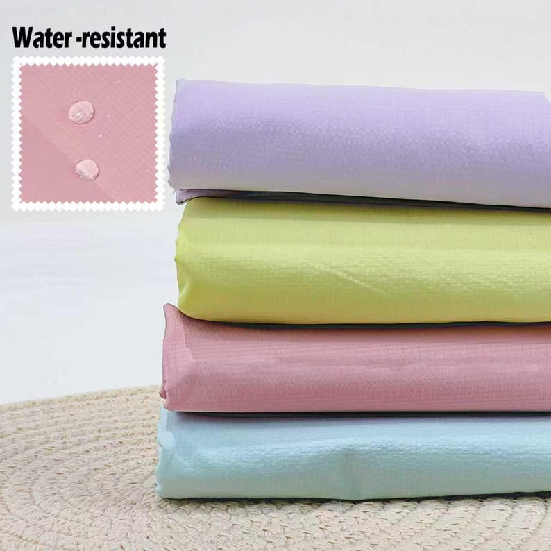 May include: A stack of four folded, water-resistant fabrics in pastel colors: lavender, yellow, pink, and light blue. The top left corner features a pink square with the words "Water-resistant" and water droplets.