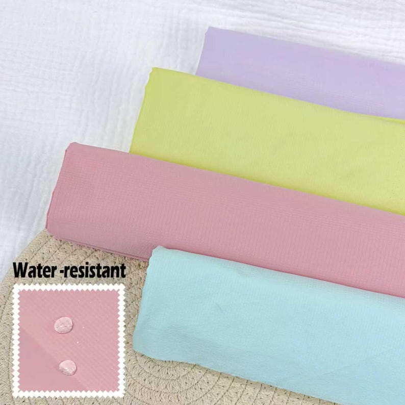 May include: Four pastel-colored fabric swatches: lavender, yellow, pink, and light blue. The pink swatch has a small square with the words "Water-resistant" and two water droplets. The fabrics are arranged diagonally, with a textured surface in the background.