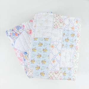 May include: A folded patchwork quilt featuring a variety of floral patterns in pastel colors. The quilt has a mix of blue, pink, yellow, and white squares, with a quilted texture. The overall design is a mix of floral and geometric patterns.