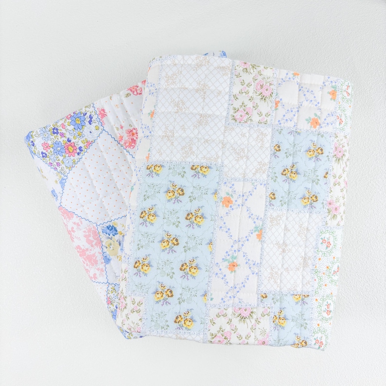 May include: A folded patchwork quilt featuring a variety of floral patterns in pastel colors. The quilt has a mix of blue, pink, yellow, and white squares, with a quilted texture. The overall design is a mix of floral and geometric patterns.
