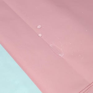 May include: Close-up of a pink fabric with a textured surface. Several water droplets are visible on the surface, creating a contrast with the solid color. The fabric is folded, revealing a light blue edge.