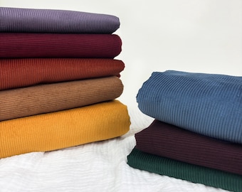 May include: Stacks of colorful corduroy fabric in shades of purple, burgundy, rust, brown, and yellow. Additional stacks of blue, burgundy, and green corduroy are also visible. The fabric is arranged on a white, textured surface.