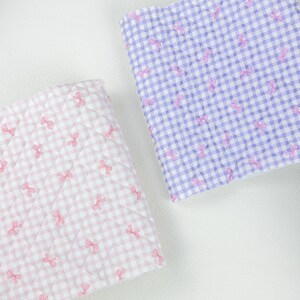May include: Two quilted fabric squares. One is pink and white gingham with pink bow accents. The other is purple and white gingham with pink bow accents. The fabric is quilted with a diamond pattern.