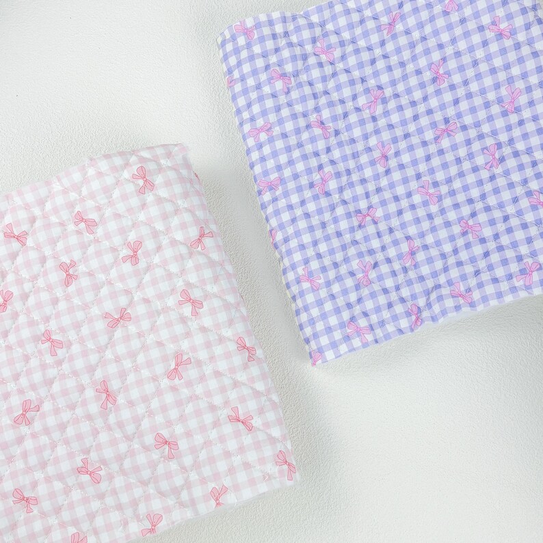 May include: Two quilted fabric squares. One is pink and white gingham with pink bow accents. The other is purple and white gingham with pink bow accents. The fabric is quilted with a diamond pattern.