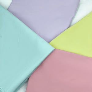May include: An overhead shot of four fabric swatches in pastel colors. The colors are light blue, lavender, pale yellow, and pink. The fabric is smooth and appears to be a lightweight material, possibly cotton or linen. The swatches are arranged in a geometric pattern.