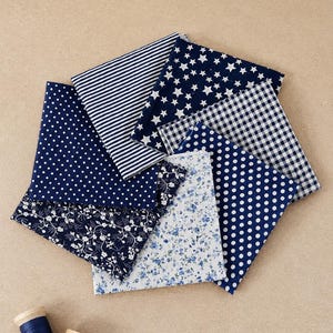 Peut inclure: Un ensemble de sept carrés de tissu disposés en éventail, présentant des motifs bleu marine et blancs incluant des rayures, des étoiles, des pois, du vichy et des fleurs, accompagnés de deux bobines de fil en bois bleu marine et blanc.