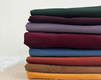 May include: A stack of eight folded pieces of ribbed fabric in various colors, including dark green, deep purple, light purple, burgundy, blue, rust orange, brown, and yellow, arranged in a vertical pile on a textured surface.