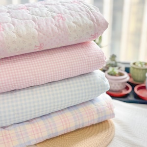 May include: A stack of four pastel-coloured quilted blankets. The top blanket is white with pink bows and floral patterns. The second blanket is pink gingham, followed by a blue gingham and a plaid blanket in pastel colours. The blankets are stacked on top of a straw hat.