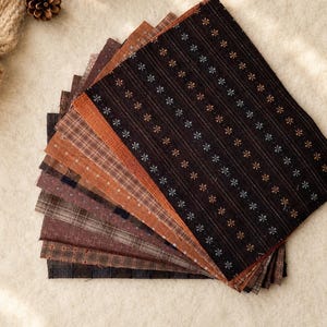 May include: A stack of rectangular fabric swatches in various shades of brown, rust, and black. The top piece is dark brown with vertical stripes and small floral patterns. Other layers feature plaid, checkered, and solid patterns in earthy tones.