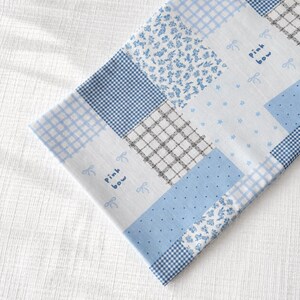 May include: A patchwork textile with a mix of blue and white patterns, such as gingham, floral, and dotted designs. The words "Pink bow" are written in cursive. The fabric is folded, showing a glimpse of the underside.