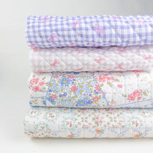 Peut inclure: Une pile de quatre couvertures matelassées. La couverture supérieure est un motif vichy violet clair avec des nœuds roses. La deuxième couverture est rose vichy avec des nœuds roses. La troisième couverture a un motif floral avec des fleurs bleues, roses et blanches. La couverture inférieure a un motif floral.
