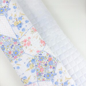May include: A rolled-up white quilted fabric with a floral patchwork design. The patchwork features squares with blue, pink, and yellow flowers on a white background. The other fabric is a solid white with a diamond pattern.