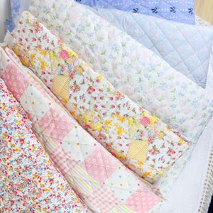 May include: A collection of colorful, quilted fabric items, including pillows and blankets. The fabrics feature floral patterns in various colors, including pink, yellow, and blue. Some items have a patchwork design, while others have a quilted diamond pattern.