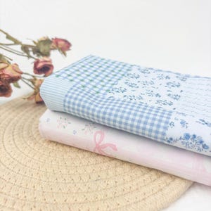 May include: Two folded fabric pieces with floral and gingham patterns. The top piece features a blue and white gingham check and floral design. The bottom piece is pink with a bow and floral pattern. The fabrics are stacked on a beige woven mat.