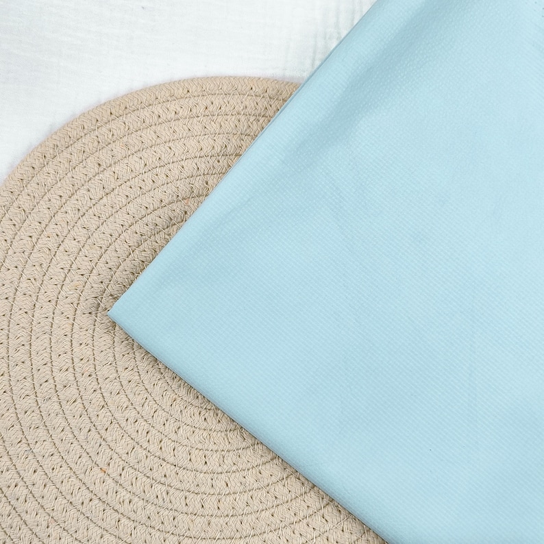 May include: A light blue fabric is draped over a beige woven placemat. The fabric has a subtle ribbed texture. The placemat is round with a detailed woven pattern. A white fabric is visible in the background.