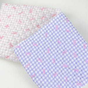 May include: Two quilted fabric squares, one pink and one lavender, feature a gingham pattern with pink bow accents. The fabric has a soft, padded appearance, suggesting a baby blanket or similar item.