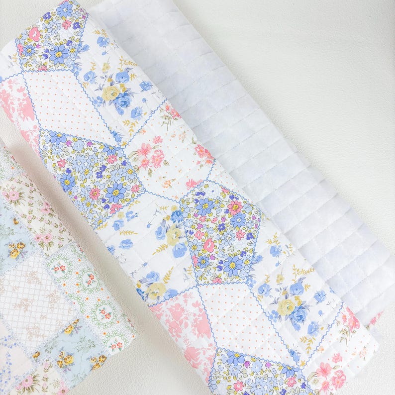 May include: A folded patchwork quilt with floral patterns in pink, blue, and yellow on a white background. The quilt is made of squares and rectangles, with a quilted texture. A second white, quilted blanket is also visible.