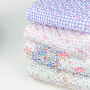 May include: A stack of four quilted blankets in pastel colors. The top blanket is lavender gingham with pink bows. The second is pink gingham with pink bows. The third has a floral pattern with blue and pink flowers. The bottom blanket has a floral pattern.
