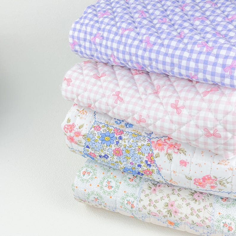 May include: A stack of four quilted blankets in pastel colors. The top blanket is lavender gingham with pink bows. The second is pink gingham with pink bows. The third has a floral pattern with blue and pink flowers. The bottom blanket has a floral pattern.