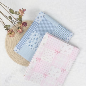 May include: Two folded fabric squares with patchwork patterns. One is light blue with floral and gingham designs, the other is pink with hearts and bows. A straw hat and dried roses are in the background.