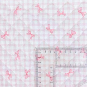 May include: Close-up of a quilted fabric with a pink and white gingham pattern. Scattered across the fabric are pink bow illustrations. A ruler is placed on the fabric, showing measurements in inches.