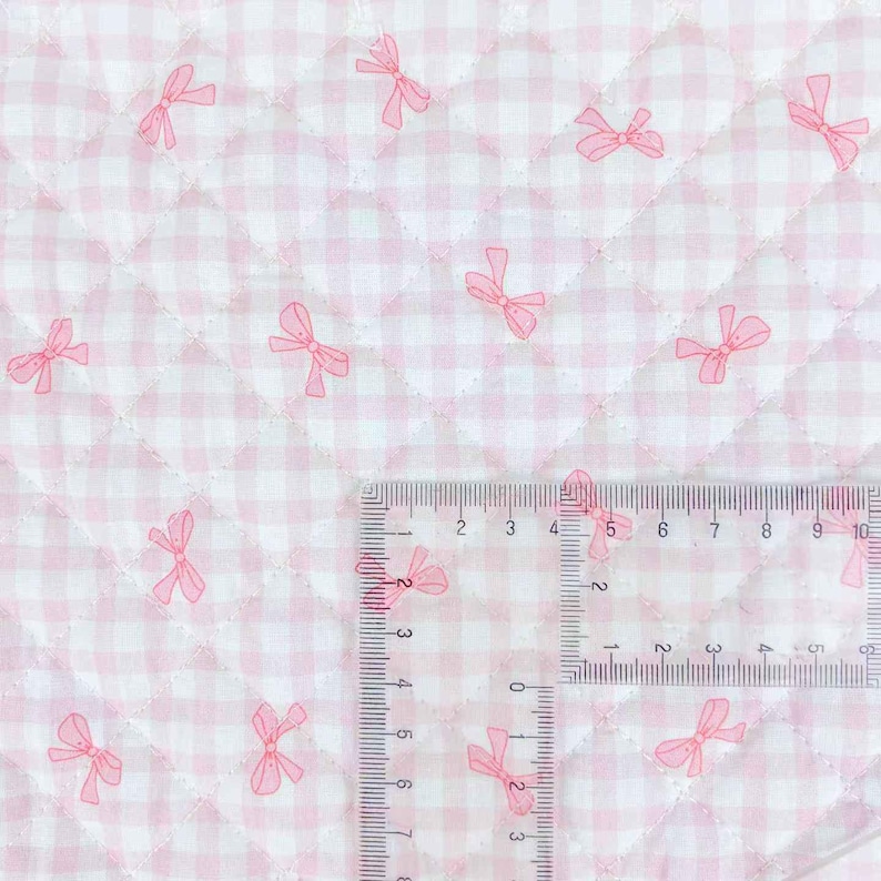 May include: Close-up of a quilted fabric with a pink and white gingham pattern. Scattered across the fabric are pink bow illustrations. A ruler is placed on the fabric, showing measurements in inches.