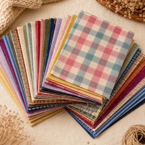May include: A stack of rectangular fabric swatches arranged in a fan shape. The collection features various patterns including gingham, stripes, and solid colors in shades of pink, blue, green, yellow, purple, and red, all with a visible woven texture.