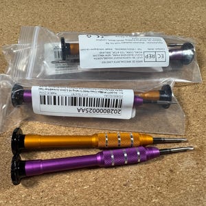 May include: An assortment of precision screwdrivers, some in clear plastic packaging. Two screwdrivers are visible, one gold and one purple, with black tips and textured handles. The background is a textured, light brown surface.