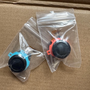 Może przedstawiać: Dwie części zamienne do joysticka, jedna niebieska i jedna czerwona, każda z czarnym wierzchem i zapakowana w przezroczyste plastikowe woreczki. Części są przeznaczone do kontrolera konsoli do gier.